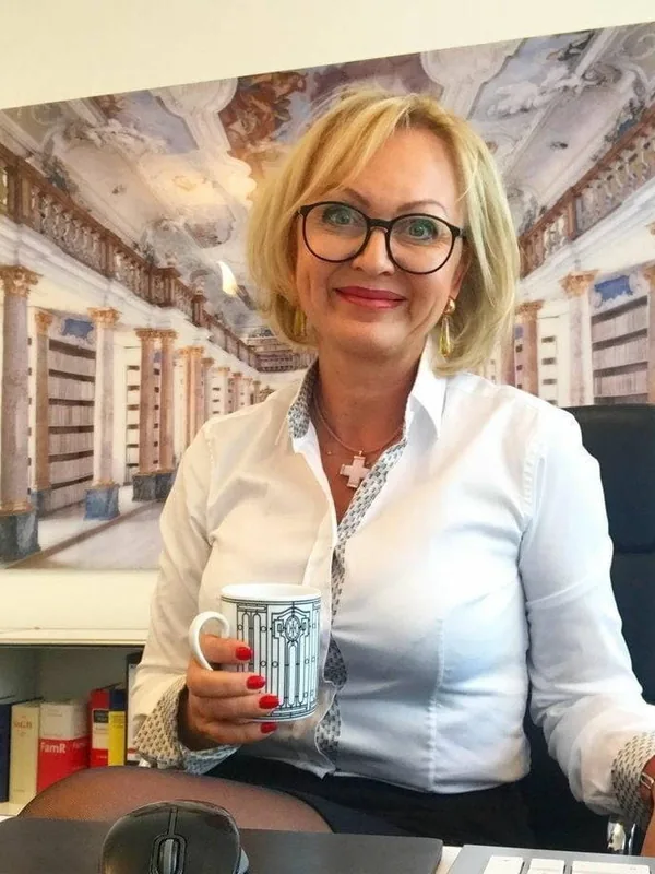 Portrait d'une personne aux cheveux blonds courts, lunettes, chemise blanche, tenant une tasse, devant un tableau de bibliothèque baroque.