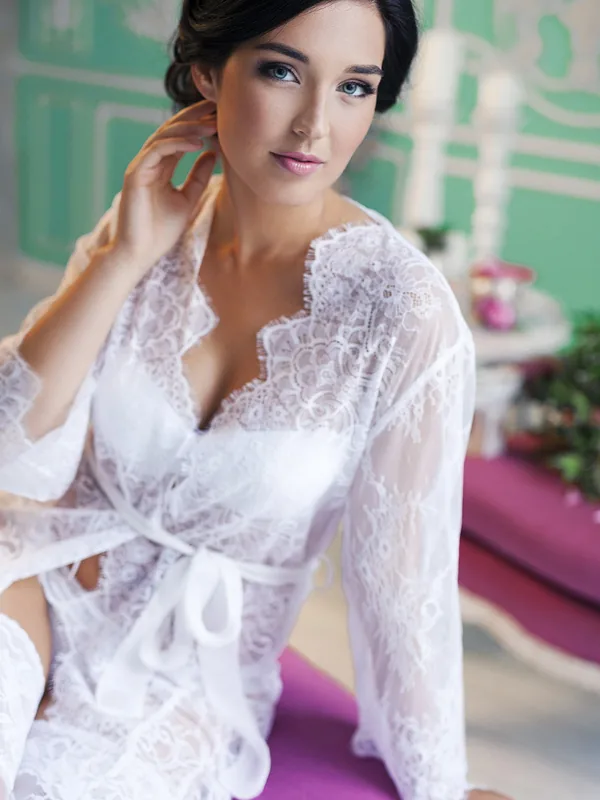 Femme aux cheveux noirs portant un peignoir blanc en dentelle, assise sur un banc violet, regardant l'objectif.