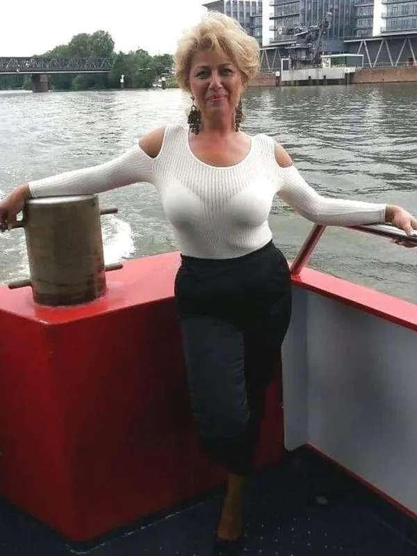 Personne aux cheveux blonds sur un bateau rouge, vêtue d'un haut blanc et d'une jupe noire, devant un fleuve et des bâtiments urbains.