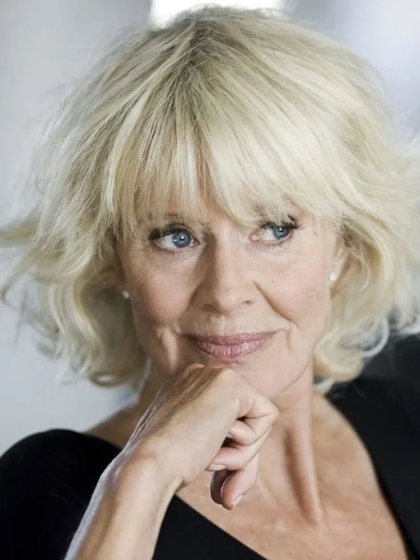 Portrait d'une femme aux cheveux blonds courts, regard bleu, sourire discret, main sous le menton, vêtue de noir.