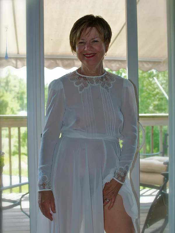 Personne en robe blanche transparente avec dentelle, souriant devant une terrasse vitrée, lumière naturelle.