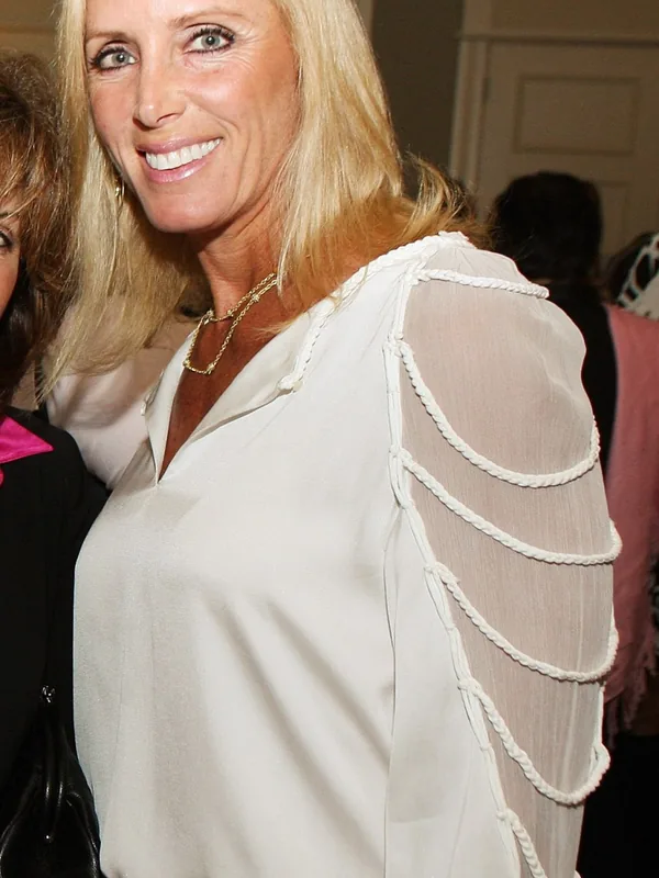 Portrait en buste d'une personne aux cheveux blonds, souriante, portant une blouse blanche à manches transparentes et une ceinture chaîne
