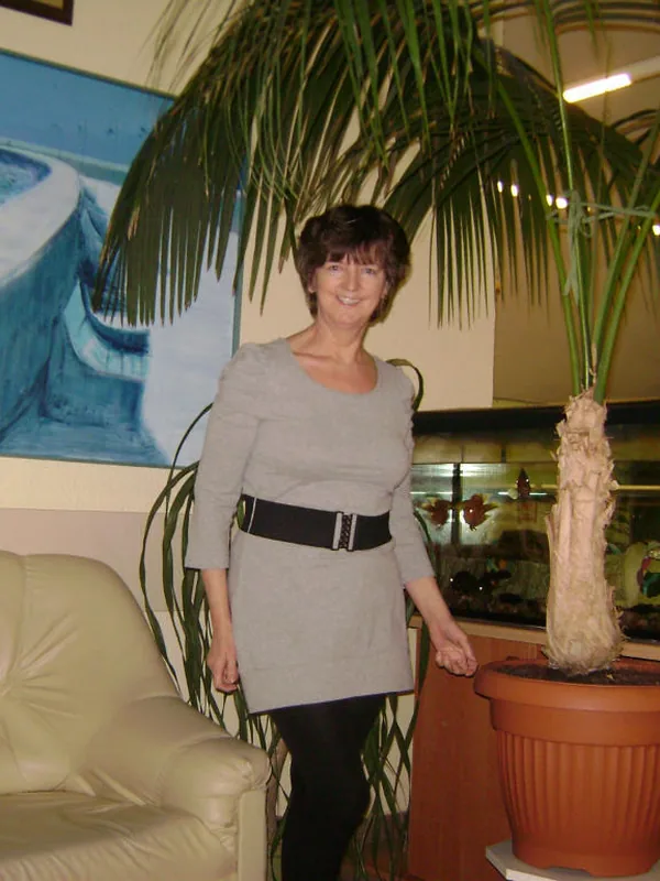 Personne souriante en robe grise près d'une plante et d'un aquarium, dans un salon avec canapé et tableau marin