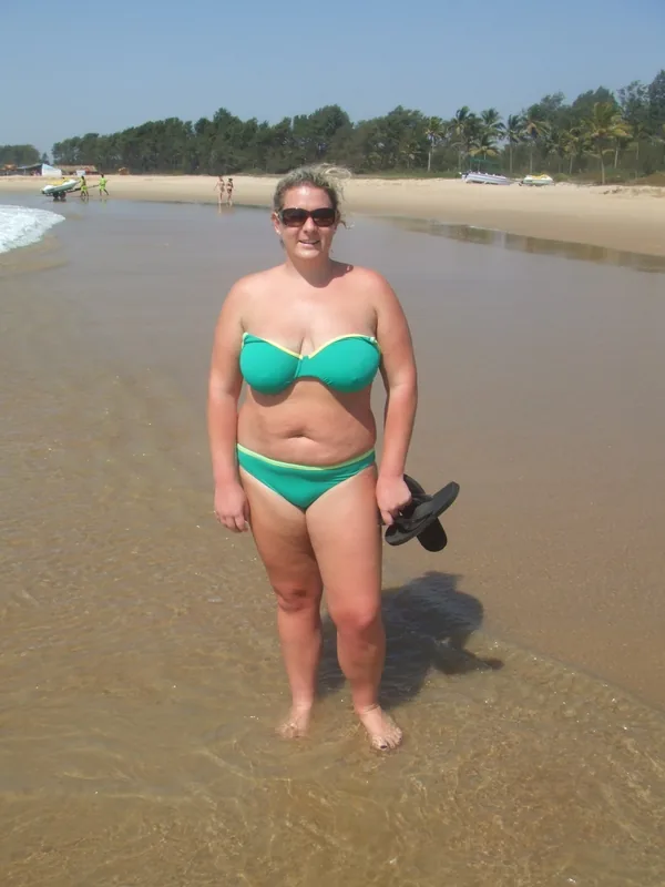 Personne en maillot vert à la plage, pieds dans l'eau, tenant des sandales, sous un ciel clair.
