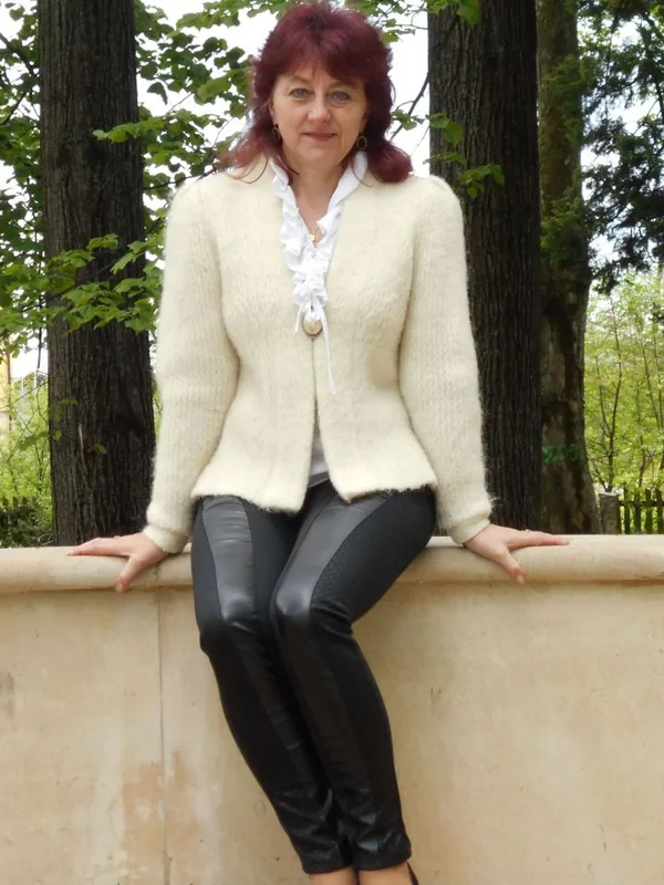 Personne aux cheveux roux portant une veste blanche et des pantalons noirs, appuyée sur un mur dans un parc arboré.