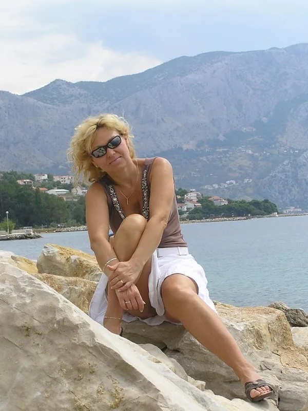 Personne aux cheveux blonds assise sur des rochers près de la mer, avec des montagnes en arrière-plan