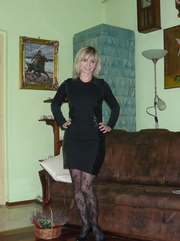Personne en robe noire et collants à motifs, posant dans un salon avec canapé en cuir et décor mural