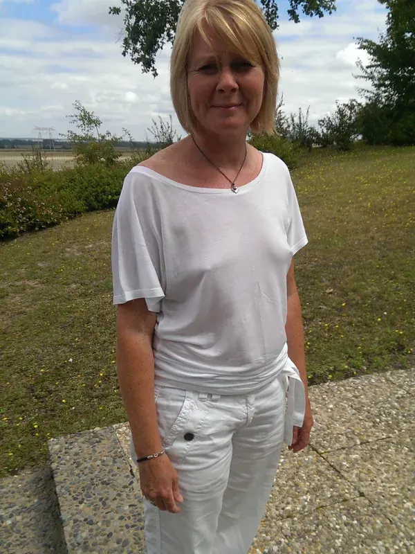 Personne en tenue blanche debout sur un trottoir en plein air, sous un ciel nuageux, entourée de verdure.