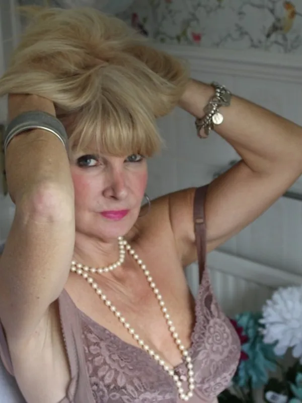 Portrait de femme aux cheveux blonds, vêtue de dentelle et de perles, les mains dans les cheveux, devant un mur décoré