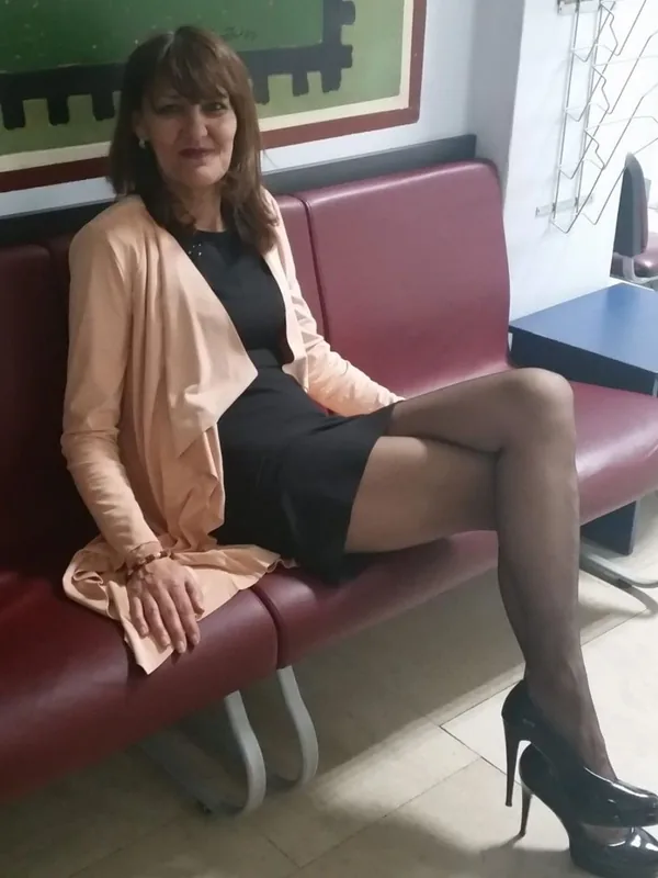 Personne assise sur un banc rouge, robe noire, veste beige, talons hauts, regard vers l'objectif