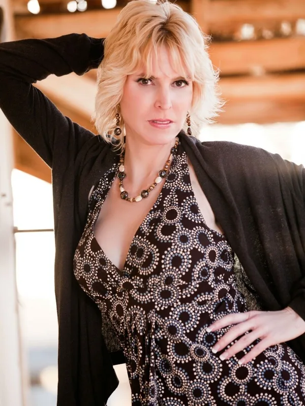 Portrait de femme blonde en robe à motifs, cardigan noir, bijoux, main sur la hanche, regard direct, lumière douce