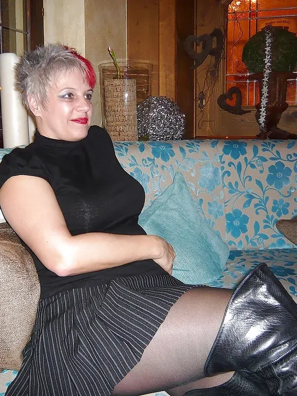 Femme aux cheveux courts gris et rouge, vêtue de noir, assise sur un canapé fleuri avec des bottes en cuir.