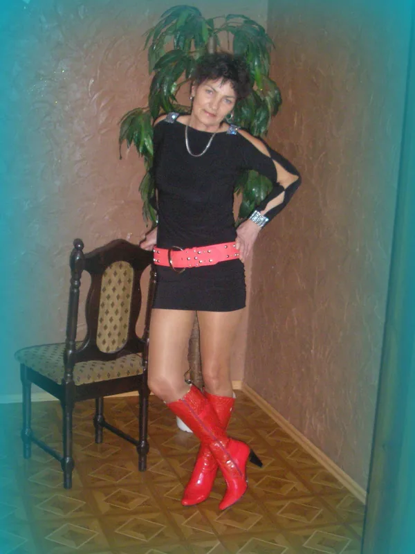 Personne en tenue noire, ceinture rose, bottes rouges, posant près d'une chaise et d'une plante
