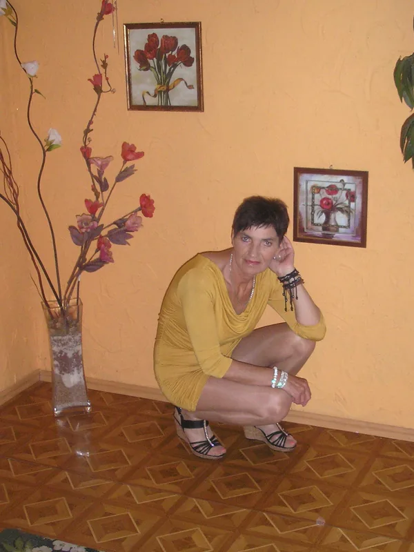 Personne accroupie en robe jaune, sur parquet, devant mur orange avec tableaux floraux et vase décoratif