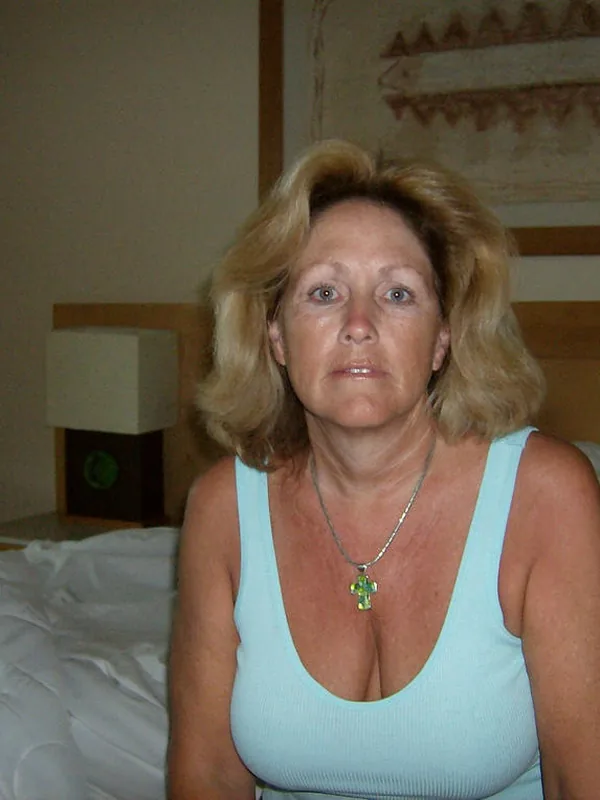 Personne aux cheveux blonds assise sur un lit, portant un débardeur bleu clair et une croix verte, dans une chambre éclairée doucement.